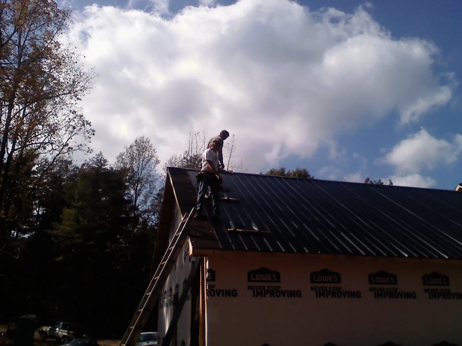 Roofer fastening dark metal panels high against a cloudy sky, from a Roof Inspection job in Houlton